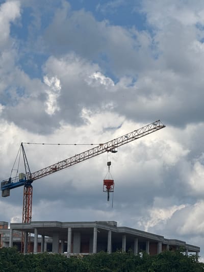 A daytime photograph captures a construction scene dominated by a large tower crane set against a cloudy sky. The image is oriented vertically (rotated 90 degrees clockwise), but describing it as if correctly oriented, the crane extends from the lower-left to the upper-right.
The main subject is a towering construction crane, featuring a prominent orange lattice jib and a blue support structure. A bright orange concrete pouring bucket is suspended by cables from the crane's jib, hanging in mid-air and indicating ongoing construction work. No operators or other people are visible on or around the crane.
To the right side of the frame, a multi-story concrete building structure is under construction, its grey pillars and floor slabs exposed, showcasing its unfinished state. Dense green foliage, likely trees or bushes, lines the far-right edge of the image, partially obscuring the building.
The sky occupies a significant portion of the background, filled with a dynamic mix of bright white and darker grey clouds. Patches of clear blue sky are visible between the clouds, suggesting a partly overcast day. The lighting is bright and diffused, indicating daytime, possibly late morning or afternoon. There are no specific location cues, visible text, or discernible emotions in the image.