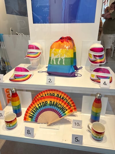 This is an indoor product display of rainbow-themed merchandise, likely a retail store or market stall. The items are arranged on white shelves. In the foreground, there are two rainbow-colored fans, a rainbow-patterned water bottle, and a rainbow-patterned mug. A price tag of "8€" is visible near the mug. Further back, a larger rainbow drawstring bag with graphics is displayed. To the left and right of the bag are rainbow-patterned visors on mannequins, with price tags of "2€" and "4€" respectively. On the far left, partially visible, are umbrellas with blue handles. In the background, a large screen displays text and imagery, and a person is holding a baby. The overall aesthetic suggests items related to LGBTQ+ pride. There is no visible indication of the city of Orihuela, Spain, within the image or the provided crops.