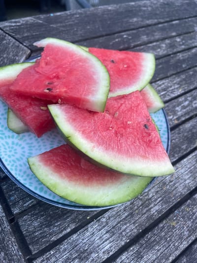 A plate of watermelon slices sits on a weathered wooden outdoor table. The watermelon slices are triangular in shape, with a vibrant red interior and a pale green rind. Some slices have visible black seeds. The plate has a light blue and white patterned design. The background shows the texture of the wooden table, which appears to be outdoors. There are no people visible in the image. The scene is captured in Maidstone, United Kingdom, suggesting an outdoor setting like a garden or patio.