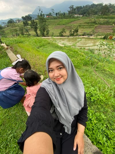 The image depicts a selfie taken in a rural landscape near Penanggungan, Indonesia. In the foreground, a woman wearing a grey hijab and black long-sleeved top smiles directly at the camera, holding the device herself. To her left, two young children are partially visible, both with their backs to the camera and bending down, interacting with something on the grassy ground. One child wears a pink shirt and blue overalls, while the other wears a pink dress. The immediate environment is characterized by lush green grass and foliage. In the background, expansive terraced rice paddies, some partially flooded and reflecting the sky, stretch across rolling hills. Distant mountains are visible under an overcast sky, with scattered trees and a few structures interspersed within the landscape. The woman appears to be seated on a concrete ledge or low wall.