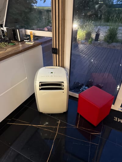 The image depicts an interior scene, likely a modern living space, with a large window or glass door offering a view of an outdoor area. Inside, a white portable air conditioning unit stands on a black, glossy tiled floor with gold veining. To the right of the air conditioner is a bright red cube-shaped ottoman. In the background, through the glass, an outdoor wooden deck is visible, along with greenery and what appears to be a rock sculpture. On a wooden countertop to the left, a toaster and a tube of lotion are present. The overall impression is one of a contemporary, possibly minimalist, home environment. The image does not contain any explicit information that would allow for the specific identification of the location as Vol'noe, Russia.