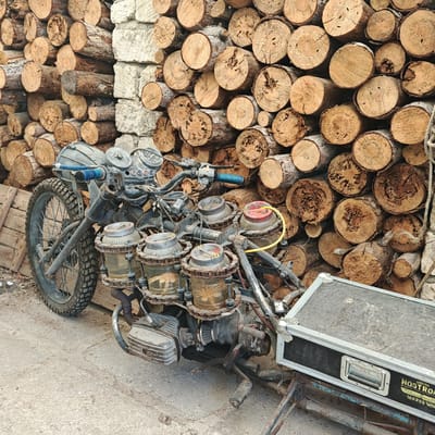 The image depicts a heavily customized, vintage-style motorcycle positioned outdoors in Останинское сельское поселение, Ukraine. The motorcycle, primarily dark in color and exhibiting significant wear and rust, is viewed from a slightly elevated angle.
The front of the vehicle features a single spoked wheel fitted with a knobby tire, suggesting off-road capability. Handlebars are visible, equipped with what appears to be a round headlight, two main circular gauges (likely a speedometer and tachometer), and several smaller indicator lights. Blue grip tape is present on the left handlebar.
The mid-section of the motorcycle showcases a highly unconventional engine design or modification. Six clear glass jars, each secured with chains and metal bands, are vertically mounted in pairs along the frame, potentially serving as decorative elements, auxiliary fuel tanks, or part of a unique intake system. One jar is topped with a red cap and has a yellow hose connected to it. An exposed air filter is visible near the engine block, which features prominent cooling fins characteristic of an older, air-cooled unit. Footpegs are present on both sides.
Attached to the rear of the motorcycle is a black equipment case, resembling a flight case, with metal corner protectors and a silver handle. The side of the case bears the inscription "USCSS NOSTROMO" above the number "180286."
The background consists of a tall, tightly stacked wall of cut logs, primarily light brown and tan, showing their tree rings and bark texture. A section of a rough, light-colored stone wall is interspersed within the log stack on the left side of the frame. The motorcycle rests on a flat, light gray concrete or paved surface. No individuals are visible in the scene.