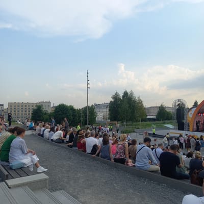 The image shows an outdoor event with a large audience seated on tiered benches. The audience is diverse in age and gender, and many are casually dressed. They are facing a stage area where a band is performing, illuminated by stage lights. The background features trees, buildings, and a cloudy sky. The location is the Путрышкаўскі сельскі Савет in Belarus, suggesting a local festival or concert.