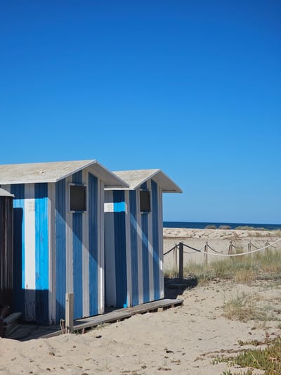 The image shows a row of beach huts at Deveses beach in Spain, under a clear blue sky. The huts are painted with alternating vertical blue and white stripes, and have white roofs. Two of the huts are fully visible, with windows and wooden boardwalks leading up to their entrances. The sand is light brown and textured, with some sparse vegetation growing on it. A rope barrier with wooden posts separates the beach huts from the dunes and the sea in the background. The water is dark blue, and a line of rocks is visible along the shoreline. There are no people or animals visible in the scene.