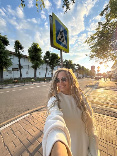 A young woman with long, wavy blonde hair and round sunglasses is taking a selfie outdoors, smiling at the camera. She is wearing a light-colored sweatshirt. The image is taken from a low angle, showing a bright, sunny scene during what appears to be golden hour, with warm light and long shadows.
In the foreground, a prominent blue and yellow pedestrian crossing sign is visible to the left of the woman. Behind her, a paved sidewalk with brick-like patterns extends alongside an asphalt road. Across the road, there is a distinctive long, white wall featuring several arch-shaped niches. This wall is lined with trees and a black metal fence. A brown directional sign near the wall, partially visible, indicates "БЛАГОВЕЩЕНСКИЙ" (Annunciation), likely part of a street or area name. In the distance, other buildings and street signs, including a prohibitory sign with a red circle and diagonal line, can be seen along the street. The sky above is blue with scattered white clouds. The overall setting is an urban or historical environment, consistent with Заволжье, Russia. The woman appears happy and relaxed.