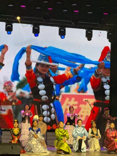 This image captures a vibrant cultural performance taking place indoors on a stage, likely in Guta, China. A large digital screen serves as a dynamic backdrop, displaying an image or video of performers in traditional attire, one actively manipulating long blue sashes or ribbons.
On the physical stage, a group of individuals, primarily women, are arranged. They are dressed in a variety of elaborate, colorful traditional or folk costumes, featuring different styles, patterns, and headwear. Some performers are seated or kneeling on the right side of the stage, wearing dresses in shades of maroon, white with intricate patterns, light blue, lime green, and black with gold embroidery. Their expressions appear composed and focused. A few male figures are visible in the background or behind the seated women. Microphone stands are positioned in front of some performers, suggesting a vocal or musical element to the show.
The left side of the frame reveals stage lighting equipment, including spotlights mounted on a truss structure, emitting blue and pink light, illuminating the stage. Other stage equipment, possibly speakers, is visible on the far right. The overall ambiance suggests an evening or nighttime event, characterized by controlled stage lighting. No discernible text is visible on the screen or elsewhere in the image.