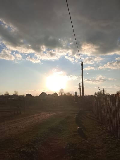 The image shows a rural scene in Высокиничи, Russia, at sunset. The sun is positioned low on the horizon, partially obscured by clouds, casting a bright glow. Several houses are silhouetted against the sunlit sky. A dirt road stretches into the distance, leading towards the houses. A wooden fence runs along the right side of the frame, and utility poles with wires are visible. The foreground consists of grassy areas.