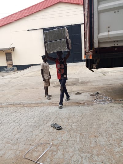 In Pasere, Nigeria, a man in a "Bulls 23" jersey carries two large, wrapped rectangular objects on his head, heading towards a shipping container attached to a truck. Another man, wearing a t-shirt and shorts, stands nearby watching. A pair of sandals lies discarded on the concrete ground, along with a pile of white strapping material and a long piece of strapping. In the background, there is a beige building with a red roof and a large metal door.