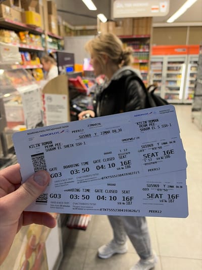 The image depicts an indoor retail environment, consistent with a convenience store or mini-market within an airport terminal. The location is not in Boldino, Russia, as the visible boarding passes indicate a flight route from Perm, Russia (PEE) to Sharm El Sheikh, Egypt (SSH-1).

In the foreground, a light-skinned left hand holds two white Aeroflot boarding passes. The thumb, index, and middle fingers are visible, with short, unpolished nails. The boarding passes are for passenger KILIN ROMAN on flight SU5969, departing on March 27th, with a boarding time of 03:50 at Gate G03, for seats 16E and 16F. The destination is Sharm El Sheikh, originating from Perm. Each pass displays a barcode, "HAVE A NICE TRIP!" in English, and "СЧАСТЛИВОГО ПУТИ!" in Russian.

In the mid-ground, a woman with blonde hair tied back in a bun is seen from behind, facing away from the camera and slightly to the right. She wears a dark (likely black) jacket with a grey hood visible at the neckline, light-colored pants, and white sneakers. She appears to be standing or moving in an aisle.

The background features extensive retail shelving units stocked with various consumer goods. On the left, shelves hold packaged snacks (e.g., chips, candies) and other items, with some products in cardboard boxes stacked above. Further back on the left, a blurry figure with light hair, wearing a light-colored top, is partially visible behind a counter or service area, possibly a checkout. On the far right, a tall, clear-doored commercial refrigerator is visible, containing bottled items. Overhead fluorescent or LED strip lighting illuminates the entire scene. The floor is light-colored.