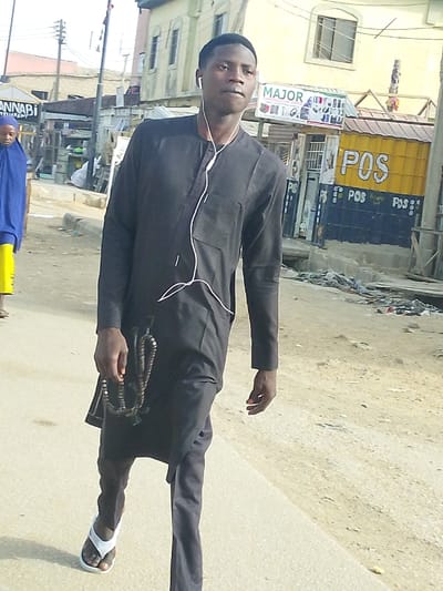 An image captured in Garun Gawa, Nigeria, depicts an outdoor urban street scene under bright daylight. In the foreground, a young adult male with dark skin and short dark hair walks towards the viewer's left. He wears a black long-sleeved traditional tunic over black trousers and white flip-flops. White wired earphones are in his ears, with the wire draped across his chest. He holds a string of dark brown prayer beads in his left hand. His gaze is directed slightly to his right.
In the mid-ground, to the left, a female figure is partially visible, walking away from the camera. She wears a blue head covering (hijab) and a yellow garment.
The background features various commercial structures and buildings. To the man's right, a yellow and blue corrugated metal kiosk displays "POS" in large lettering. Above it, a white sign with "MAJOR" in red text advertises electronics. Behind these structures is a light-colored multi-story building with windows. Utility poles and wires are visible against a bright sky. The street surface appears to be a light-colored paved road, with debris and dirt present along the right edge.