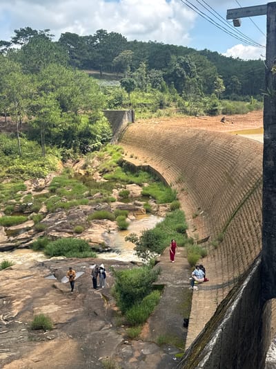 A curved concrete dam with a rough, textured surface is the dominant feature in the foreground and midground of the image, set against a backdrop of lush green hills and trees. The dam's surface is patterned with numerous small, rectangular indentations, forming a repeating grid-like design. On the flat area near the base of the dam, several people are visible. One woman in a red dress walks along a path to the left. Further to the right, a group of three people are seated on the dam's surface, one wearing a plaid shirt. On a rocky outcrop to the left of the dam, a group of three individuals stand, one holding a camera. The water below the dam flows over rocks, creating a stream with surrounding greenery. The background consists of a dense forest of pine trees covering rolling hills. Power lines stretch across the top right of the frame, connected to a concrete structure. The sky is partly cloudy with patches of blue. The scene appears to be an outdoor recreational area, possibly a tourist attraction, located in Lang Biang - Da Lat, Vietnam, given the distinctive dam architecture and surrounding natural landscape.