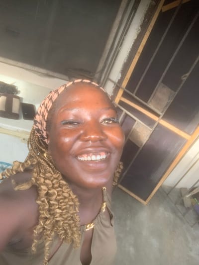 A woman with dark skin and blonde braided hair, wearing a patterned headscarf and a brown top with gold accents, smiles at the camera. Her eyes are open, and her teeth are visible. She is wearing a gold hoop earring. The background appears to be an indoor space with a tiled floor and a wall with wooden slats. The overall lighting is bright, suggesting daylight. The specific location in Eliozu, Nigeria, is not discernible from the visual information provided in the image.