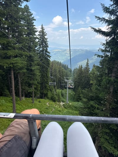 The image captures a scenic view from a ski lift traveling through a lush, forested mountain landscape in Panichishte, Bulgaria. The foreground shows the legs of two individuals seated on the ski lift, one wearing brown shorts and the other light-colored leggings. The ski lift's metallic railing runs horizontally across the bottom of the frame. In the mid-ground, several empty ski lift chairs are visible, along with a chair carrying passengers further up the slope. The surrounding environment is dominated by dense evergreen trees, with rolling hills and distant mountains stretching towards the horizon under a partly cloudy blue sky. The ski lift cables are clearly visible, connecting the chairs and extending upwards and across the frame. The ground beneath the ski lift is covered in thick green vegetation and smaller trees.