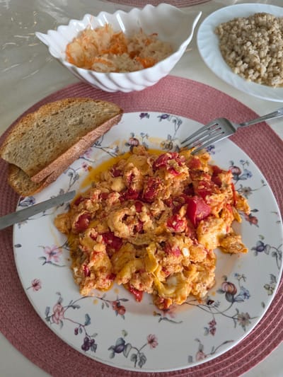 In Skolkovo Innovation Center, Russia, a close-up overhead shot captures a breakfast setting on a white table covered with a pink placemat. The focal point is a white plate adorned with floral patterns, holding a serving of scrambled eggs mixed with diced red tomatoes. Two slices of toasted bread rest to the left of the plate, with a knife positioned on the left side of the plate. A fork is placed on the right side of the plate. In the background, a white, wavy-edged bowl contains a light-colored salad, possibly sauerkraut with shredded carrots. To the right of this bowl, another white plate displays a pile of cooked barley or another grain. The scene is well-lit, highlighting the textures and colors of the food.