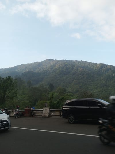 The image depicts a roadside scene against a backdrop of a forested mountain in Taunggyi, Myanmar. The sky is light blue with scattered clouds. In the foreground, a black SUV is partially visible on the right side of the road. A silver car is visible on the left. Several people are seen on the roadside, some sitting or standing near the barrier. A few motorcycles are parked nearby. The overall environment appears to be a roadside overlook or rest stop.