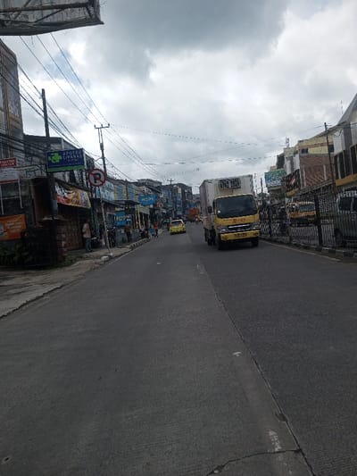 The image displays an urban street scene in Gubbisamamidi, India, under an overcast sky. A two-lane asphalt road, divided by a central seam, extends into the distance. On the left, a sidewalk fronts a continuous row of multi-story commercial buildings, featuring visible signage for "PRAKTEK DOKTER" and "APOTEK CIPANAS," along with a red circular 'No Parking' sign. A dense network of overhead wires and utility poles spans above the street. Approximately six individuals are present on the left sidewalk, some standing near storefronts, others walking. Parked motorcycles are visible near the buildings on the left. On the right side of the road, a black metal fence borders another series of commercial structures. A yellow box truck with a white refrigerated unit, bearing "JL-100" on its grille, occupies the right lane and is moving away from the viewer. Further down the road, a yellow car is also in motion, following the truck. Other distant vehicles and pedestrians are indistinct.