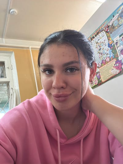 A young woman with dark hair and light brown eyes, wearing a pink hooded sweatshirt and a gold chain, is photographed indoors. Her left hand is resting on her neck, and she is looking towards the camera with a slight smile. The background includes a yellow wall, a window with white trim, and a colorful poster on the wall. The overall lighting suggests an indoor setting, possibly a classroom or personal space. There is no indication of the city of Чумна, Russia, in the image or its context.