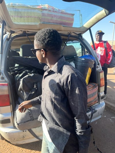 A man in a gray shirt and glasses stands near an open car trunk, filled with bags and containers. Another man, wearing a red shirt with a white skeletal design and a baseball cap, is positioned behind the car, partially visible. The trunk contains items like black duffel bags, a cylindrical container, and a striped bag. The car appears to be an older model, with visible wear and tear. The scene is bathed in sunlight, suggesting an outdoor setting.