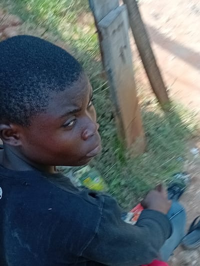 An outdoor scene in Ifite-Awka, Nigeria, depicts a young Black male, appearing to be a child or adolescent, positioned in a seated or kneeling posture. He has short, dark, closely cropped hair and directs his gaze upward and to the right. The individual wears a dark long-sleeved shirt with a visible tear on the right shoulder. A red garment is partially visible beneath his shirt, and light-colored trousers are present, with a dark shoe or slipper on his left foot. His right hand rests on an obscured item. The immediate environment consists of a grassy area transitioning to a dirt path. Two vertical wooden posts are visible in the mid-ground. Scattered debris, including a green and yellow plastic object, is present on the ground. The scene is illuminated by strong daylight.