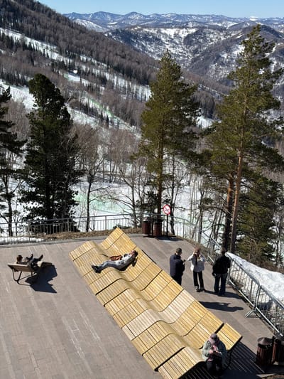 The image captures a scenic overlook in Ozernoe, Russia, with a view of snow-dusted mountains and forested slopes. A wooden platform, patterned with waves, stretches out from a paved area, serving as a seating area for visitors. Several individuals are present: one person is lying down on the wooden platform, another sits on a nearby bench with a companion, and a group of three stand near a railing, seemingly admiring the view or taking photographs. A "no smoking" sign is visible in the middle ground, near a cluster of bare trees. The overall atmosphere suggests a tranquil scene, with people enjoying the landscape on a clear day.
