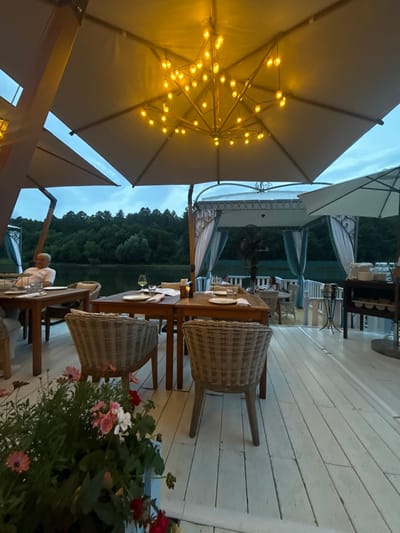 The image depicts an outdoor dining area with tables and chairs set up on a white wooden deck. In the foreground, there is a planter filled with pink and red flowers. Several tables are visible, set with plates, glasses, and napkins. In the background, there is a body of water surrounded by a dense forest. Overhead, large umbrellas provide shade, and strings of warm-toned lights are illuminated. A man in a white shirt is seated at one of the tables on the left. The overall atmosphere suggests a restaurant or resort setting by the water, likely in Zhukovka, Russia.