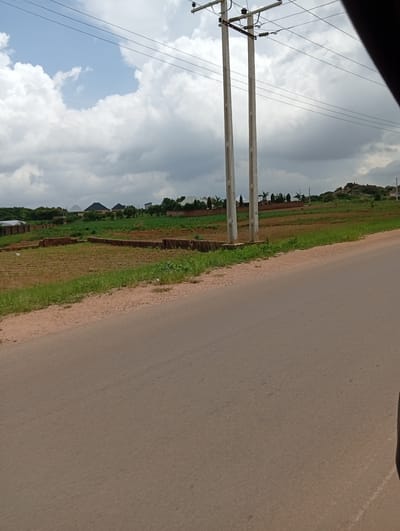 The image shows a rural or semi-rural landscape in Reifild-Ist, Nigeria. In the foreground, a paved road curves to the right, bordered by a sandy shoulder. The middle ground features cultivated fields, some with low brick walls delineating plots, and patches of green grass and vegetation. In the background, several buildings with domed and peaked roofs are visible, interspersed with trees. Two tall, concrete utility poles with crossbars and wires stand prominently in the middle ground, one slightly to the left of center and another to its right. Beyond the buildings and fields, a rocky, elevated terrain is visible on the far right, under a sky filled with scattered white and gray clouds. The scene appears to be captured from a moving vehicle, as indicated by the slight blur and the dark object encroaching on the right edge of the frame.