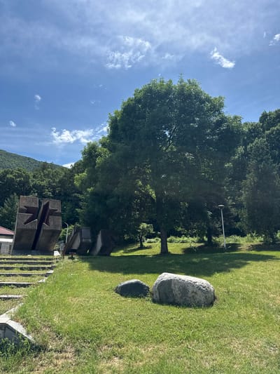 This image depicts an outdoor memorial or park area in Vetren Dol, Bulgaria, bathed in bright midday or early afternoon sunlight, evident from the strong shadows cast on the lush green grass. The sky is clear blue with scattered white, wispy clouds.
In the foreground, a large, weathered concrete monument dominates the left side. It comprises several heavy, block-like structures, two of which feature deeply carved, stylized star or cross-like shapes. A small section of a building with a reddish-orange roof is visible at the very bottom left. The ground around the monument is covered in green grass.
The midground features a sprawling green lawn where two smooth, grey boulders of differing sizes are placed. A tall, slender white pole, possibly a lamppost, stands upright among the trees. Numerous large, mature trees with dense green foliage provide shade and frame the scene, their leaves vibrant under the sun.
In the background, a dense forest covers a steep hillside or mountain, extending under the clear sky. No people or specific activities are visible, lending a tranquil and contemplative atmosphere to the scene. While there appears to be a plaque or inscription on one of the concrete monument blocks, the text is not legible due to the distance and resolution.