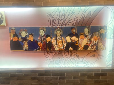 The image shows a rectangular artwork hanging on a wall. The wall is divided into two sections: a lower section made of grey brickwork and an upper section that appears to be wallpapered or painted with a light pink-beige color. The artwork depicts a group of people in what appears to be a traditional Chinese opera setting. The figures are dressed in elaborate costumes and makeup. Behind the artwork, the wallpaper features an illustration of a large, stylized phoenix design.
There is not enough information to place the image in Hailing District, China.