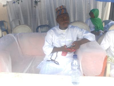 This image depicts a gathering in Ijoko, Nigeria. The central figure is a dark-skinned male, approximately 25-35 years old, seated on a peach-colored sofa covered with a translucent protective sheet. He wears a white tunic with light green embroidery and a patterned cap featuring brown, blue, and white geometric designs. He holds a silver mobile phone in his left hand and a small red fabric item in his right; dark prayer beads rest on his left arm. His gaze is directed to the right, off-camera. To his right and slightly behind him, a dark-skinned female, clad in a light-colored garment and a bright green hijab, is seated, with her hands clasped. She also looks towards the right. The background features white draped fabric adorned with blue and white artificial floral decorations, and several white and dark-colored plastic chairs. In the immediate foreground, a white surface, likely a table, holds a plastic bottle of "Aqua Rapha" brand bottled water with a blue cap. The setting suggests an event, possibly outdoors or in an open-sided tent structure.