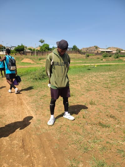 The image depicts an outdoor scene in what is identified as Barikin Jirga Summa, Nigeria. The primary subject is a young male standing in the foreground, facing downwards, and wearing a light green hoodie with the word "GALLERY" printed on it in white lettering. He also wears dark shorts over black leggings, white socks, and white athletic shoes. He has a black baseball cap on his head. A white drawstring is visible hanging from his hoodie. To the left, another young male is walking away from the camera, wearing a bright blue t-shirt, black shorts, black socks, sandals, and carrying a black backpack and a purple bag. In the background, there are several structures, including brick walls, houses with varying roof types, and some hills covered with vegetation. The ground is a mixture of dirt and sparse grass. The sky is clear and blue. The overall impression is a casual, everyday scene in a semi-rural or developing urban environment.