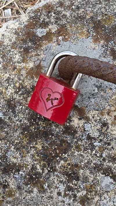 The image displays a close-up, downward-angled view of a red padlock secured to a heavily rusted metal bar or chain link, resting on a weathered, lichen-covered stone surface. The padlock body is rectangular and painted bright red, featuring a deeply embossed heart shape on its visible face. Within the heart's depression, a dark, irregularly shaped area of corrosion or detail is present, possibly a keyhole or a decorative element. The padlock's shackle is metallic silver and appears polished. The rusted metal object is thick, cylindrical, and exhibits significant surface oxidation, ranging in color from dark brown to reddish-orange. The underlying surface is a rough, mottled stone, covered extensively with various types of lichen, displaying patches of white, grey, brown, and pale green. In the upper left quadrant, dry, light-colored vegetation is visible, suggesting an outdoor setting. The scene is located in Passau, Germany.