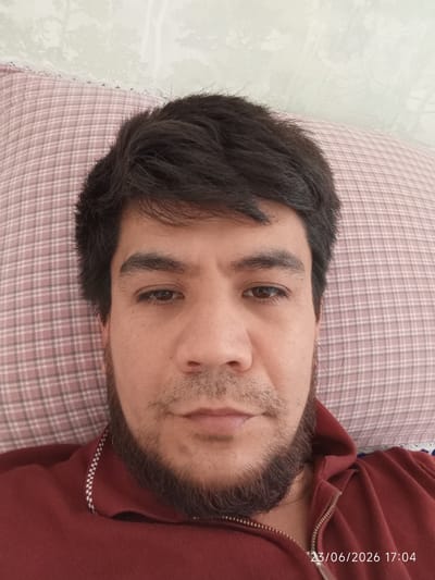 The media file shows a close-up, overhead selfie of a man lying down on a pillow. The man has dark hair, dark eyebrows, and a beard. He is wearing a dark red collared shirt, possibly a hoodie, with a zipper. The pillow he is lying on has a pink and white checkered pattern and is placed against a wall with a light-colored, textured wallpaper featuring a floral or tree-like pattern. The scene appears to be indoors, likely in a bedroom. The lighting suggests it is daytime, and there are no visible indications of weather. The man's expression is neutral. The visible text in the lower right corner reads "23/06/2026 17:04", indicating the date and time the photo was taken. The location context provided is Eshanguzar, Uzbekistan.