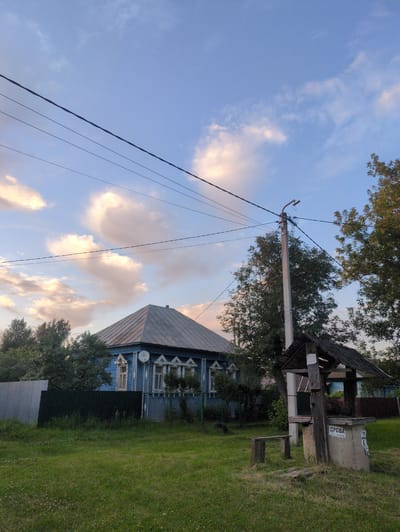 This image depicts a rural scene in Velikovo, Russia. The sky is a pale blue with scattered, wispy clouds tinged with pink and orange, suggesting it is either dawn or dusk. A prominent telephone pole with multiple wires extends upwards diagonally across the frame, with some wires leading to a small, rustic wooden structure resembling a well or pump house. This structure has a dark, sloped roof and is positioned on a patch of concrete surrounded by vibrant green grass.
Adjacent to the well structure, and further into the scene, is a traditional blue wooden house. The house features white trim around its windows, some of which have decorative triangular elements above them. A satellite dish is visible on the roof of the house. Trees with full green foliage are interspersed throughout the landscape, casting shadows and adding to the natural setting. The overall mood is peaceful and tranquil, evoking a sense of rural life.
There is some visible text on a white label attached to the concrete base of the well structure. The most prominent word appears to be "ДРОВА" (Drova), which translates to "firewood" in Russian. Below this, a phone number is partially visible. The grass is well-maintained, indicating some level of upkeep, while the wooden structures and house suggest an older, established dwelling.