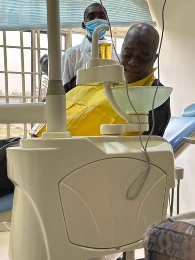 The image depicts a dental examination in progress, possibly in Tudun Nupawa, Nigeria. An older Black male patient lies in a light blue dental chair, wearing a yellow protective covering. A dental professional, wearing a white coat and a blue surgical mask, stands nearby. Dental equipment, including a white examination light and the dental unit, is prominently featured. A window with bars is visible in the background.