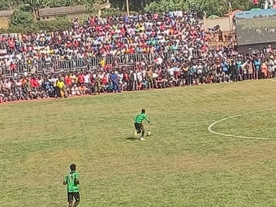 This image captures an outdoor football (soccer) match or practice session in Siaya, Kenya, on a bright, sunny day. The primary subjects are the numerous spectators and two football players on the pitch.
A large crowd, consisting of hundreds of people, fills the tiered stands and terraces along one side of the football field. The spectators are dressed in various casual clothing, creating a colorful mosaic. They appear to be actively watching the game, indicating an engaged and lively atmosphere.
On the green grass pitch, two players are visible. In the mid-ground, a player wearing a green patterned jersey and dark shorts is seen dribbling a white football. His body language suggests he is actively moving with the ball. Another player, also in a green patterned jersey and dark shorts, is running in the foreground, facing away from the camera. Part of a white semicircular line, likely a marking for the penalty area or center circle, is visible on the right side of the field.
The strong, clear lighting suggests it is either midday or early afternoon, with bright and clear weather conditions. Behind the crowded stands, some buildings and dense green foliage, possibly trees or shrubs, are visible, providing a glimpse of the surrounding environment. There is no discernible text visible on the players' jerseys, stadium structures, or any advertising. The image effectively conveys the dynamic energy of a community sporting event.