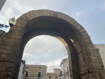 The image displays a large, ancient stone archway, likely a Roman structure, situated in Mérida, Spain. The arch, constructed from substantial, irregularly cut grey stones, dominates the foreground, framing the view beyond. An ornate black metal street lamp with a frosted glass globe is affixed to the upper left side of the arch.
Through the archway, an urban landscape unfolds under an overcast sky with scattered white clouds, suggesting a daytime scene, possibly late morning or afternoon. To the left, a white building with a brown roof and dark doors is partially visible. Further in the background, a cream-colored building with dark window frames and a small antenna on its roof can be seen. On the right, another white building with blue decorative elements, possibly balconies, is partially obscured by the arch's right support.
The overall impression is one of historical permanence integrated into a modern town. No people are visible, and no specific activity is taking place. The weather is bright but overcast, creating a soft, diffused light. No visible text is present in the image.