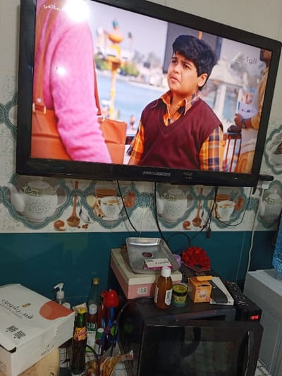 This image depicts an indoor scene with a wall-mounted television above a cluttered counter. The television, a black flat-screen model, displays a program featuring a young boy with dark hair and a round face, wearing a plaid shirt and a burgundy V-neck sweater vest, looking upwards with a serious expression. To his left, the torso of an individual in a pink top with a brown shoulder bag is partially visible. The background of the TV program shows an outdoor waterfront, including a body of water, distant buildings, and a decorative pole structure, consistent with the Shatt al-Arab Corniche in Basra, Iraq. Arabic text "الماضي" (The Past) and "الوان" (Colors) with a 'Z' logo are visible on the screen.
Below the television, the wall is tiled in two sections: an upper section with light-colored tiles featuring repeating patterns of teapots, teacups, and coffee beans with a scalloped border, and a lower section of plain, dark teal-colored tiles. Electrical wires are visible hanging from behind the television.
A counter below the tiled wall is laden with numerous items. From left to right, these include a white cardboard box labeled "Oasis Station" with Arabic text, a clear pump-action dispenser bottle, a dark brown bottle with a red label and yellow cap, a red squeeze-top bottle, a rectangular metal tray containing a red circular object and a white label, a light pink box, a red textured decorative item, a small yellow box, a small green jar, and multiple black remote controls. A dark-colored appliance, likely a microwave, with a reflective front panel and a digital clock displaying "18:05", is also on the counter. A portion of a large blue container is visible on the far right.