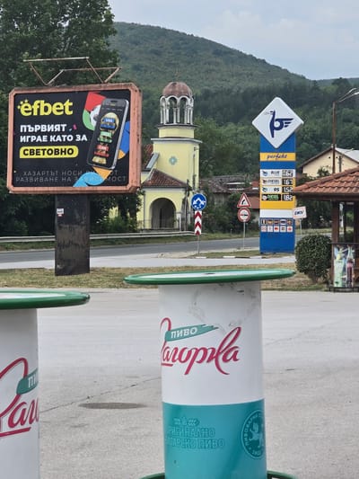 The image captures a roadside scene in Zlatna Panega, Bulgaria. In the foreground, two cylindrical structures with green rims are visible, one on the left and one on the right. The right cylinder features branding for "пиво Загорла" with a red script logo and a turquoise band at the bottom displaying wheat stalks and a circular emblem with a lion and the year "1902".
Behind the foreground structures, a billboard advertises "efbet" with the slogan "ПЪРВИЯТ ИГРАЕ КАТО ЗА СВЕТОВНО", translating to "The first plays like for the world". The billboard also includes a warning about gambling addiction. Further behind this, a yellow church with a brown dome and bell tower stands prominently. To the right of the church, a blue gas station sign displaying "perfekt" and fuel prices for "BLUE FORCE" and "DIESEL" is visible. The prices listed are 0.62 for BLUE FORCE and 1.67 for DIESEL PRO FORCE, 1.52 for 95 DIESEL, and 1.82 for DIESEL GREEN FORCE.
Several road signs are present, including a blue circular sign with a white arrow, a triangular warning sign with two red circles, and a chevron sign. The background consists of a lush, tree-covered hillside under a cloudy sky. Residential buildings with tiled roofs are visible behind the gas station sign.