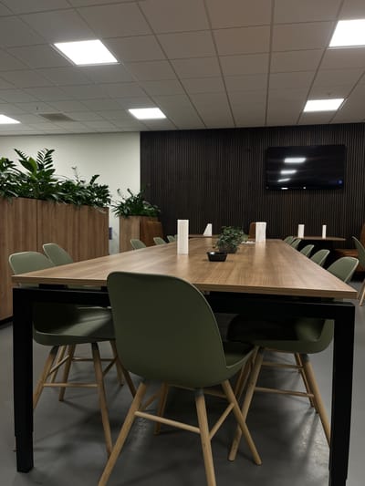 The image depicts a modern conference room. A long, light-wood conference table dominates the foreground, with several green chairs arranged around it. The chairs have wooden legs and a modern, ergonomic design. In the center of the table, there are a few decorative items, including a small plant and white cylindrical objects that appear to be candles or diffusers.

The room features a neutral color palette. The walls are white on the left, and a dark wood-paneled wall is visible on the right. A large flat-screen television is mounted on the wood-paneled wall. The ceiling is a grid of acoustic tiles with integrated LED panel lights, providing bright, even illumination.

To the left of the table, tall wooden planters filled with lush green plants add a touch of nature to the space. The floor is a uniform grey, likely polished concrete or a similar material. The overall impression is one of a clean, functional, and contemporary workspace. The image does not contain any individuals or indication of the city of Oldenzaal, Netherlands.