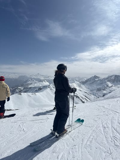 The image displays two individuals on a snow-covered mountain slope within the ski area of Le Monêtier-les-Bains, France. The foreground is dominated by a person, seen from behind and facing right, wearing a black ski helmet with goggles pushed up, a black full-body ski suit, and black gloves. This individual holds two ski poles and stands on skis, which have light blue/green bases visible. To the left, a second person is partially visible, oriented slightly away from the viewer, wearing a beige jacket, dark pants, and a red and white knitted beanie, standing on a snowboard. Both individuals are stationary, observing a vast expanse of snow-capped mountain peaks and ridges stretching across the horizon under a clear blue sky with scattered wispy clouds. The snow-covered terrain shows visible ski tracks, and prominent shadows cast on the snow indicate bright sunlight.