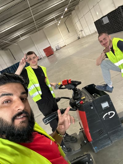 Three men are posing for a selfie in a large, empty warehouse, possibly a distribution center. The man in the foreground on the left is taking the photo, showing a portion of his face and wearing a bright yellow safety vest over a red t-shirt. He has a beard and is making a peace sign with his right hand. To his right, a second man is partially visible, also wearing a yellow safety vest over a black t-shirt and making a peace sign with his left hand. He is holding a clear plastic bottle with a yellow cap. Behind them, the third man is kneeling and smiling at the camera, wearing a yellow safety vest and dark pants. He is holding a pink object, possibly a pen. They are standing on a concrete floor. In the background, there are rows of stacked black pallets and some red storage containers. The ceiling of the warehouse has exposed beams and fluorescent lighting. The overall environment suggests an industrial or logistical setting. The man in the foreground has a black band on his left wrist. The warehouse appears to be an Amazon logistics center in Schweitenkirchen, Germany, given the branding visible on one of the pallet stacks.