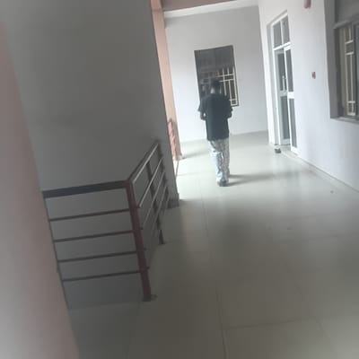 This media file captures an interior scene, likely a hallway or corridor within a building. The perspective is from a high angle, looking downwards. Dominating the frame is a person standing with their back to the camera, wearing a dark shirt and checkered pants. They appear to be looking downwards or at something on the floor.
The environment is constructed with light-colored walls and a tiled floor. There are windows visible on the walls, some with grilles, suggesting a functional rather than decorative purpose for these openings. A prominent feature in the lower right of the image is a set of railings, possibly belonging to a staircase or balcony, with reddish-brown supports and silver-colored horizontal bars. The lighting in the scene is somewhat dim, casting shadows and creating a slightly muted atmosphere.
There are no overt indicators of the time of day or weather conditions. The location context provided is Wukari, Nigeria, which might suggest a building typical of that region, though without more visual cues, specific architectural styles or cultural elements are not definitively discernible. The overall impression is one of a functional building interior with a single individual present. No visible text is present in the media.