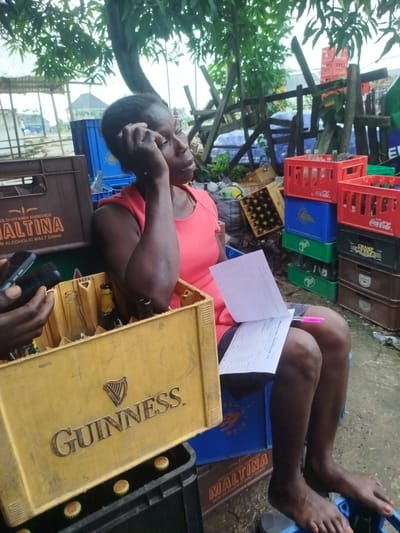 In Yenegoa, Nigeria, a woman sits amidst stacks of beer crates and bottles, seemingly overseeing operations. She is wearing a vibrant pink top and dark shorts, her posture relaxed as she holds a document and a pen. Her hand rests on her head, suggesting a moment of contemplation or perhaps fatigue. The scene is outdoors, under a leafy tree that provides some shade. The ground is wet, indicating recent rain.
Various crates and bottles are visible, including Maltina, Guinness, and Coca-Cola. Some bottles are empty, while others appear to be full or partially filled. A yellow Guinness crate is prominent in the foreground, containing several empty bottles. The presence of these beverages, along with the woman's engagement with a document, suggests a business transaction or inventory management is taking place. The background shows more crates, wooden structures, and some scattered debris, indicating a bustling but perhaps somewhat disorganized marketplace or distribution point. The overall impression is one of a hardworking individual managing a beverage distribution business in a tropical environment. The lighting suggests it might be daytime, possibly after a rain shower.