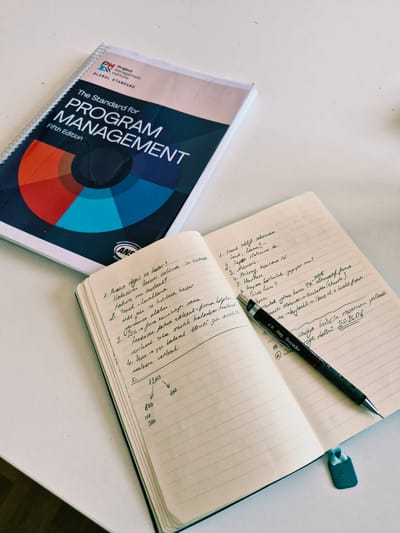 The image depicts a desk with a book titled "The Standard for Program Management, Fifth Edition" by the Project Management Institute and an open notebook with handwritten notes. A black Rotring Tikky 0.7 mechanical pencil lies on the notebook. The notebook contains a list of numbered items, some with sub-points and a simple diagram with arrows and numbers. The environment appears to be an indoor setting, with the objects placed on a flat surface. The image does not contain any information about the city of Đenovići, Montenegro.