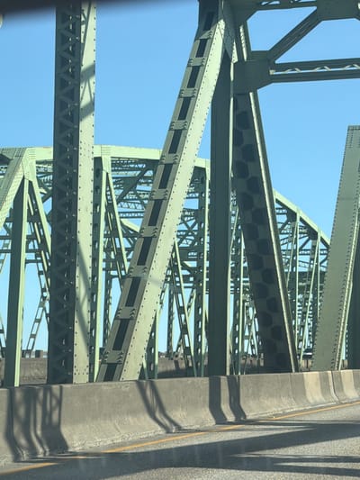 This is a close-up, angled photograph of the steel structure of a bridge, likely taken from a vehicle moving across it. The dominant subjects are the interconnected girders and beams of the bridge, painted a muted green. The sky is a clear, bright blue, suggesting a sunny day. The time of day appears to be daytime due to the clear sky and the shadows cast on the bridge structure. The scene is urban or semi-urban, given the presence of a large bridge and what appears to be a roadway on the right side of the frame, marked with a yellow line and concrete barriers. There are no people visible. The overall impression is one of industrial strength and engineering. The location is suggested to be Vancouver, USA. There is no visible text in the image.
