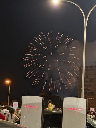 The image depicts a nighttime scene in Alicante, Spain, featuring a fireworks display over a cityscape. In the foreground, several white Tesla Supercharger stations are visible, with the "TESLA" logo illuminated in red. A crowd of people is gathered around the charging stations, some looking up at the fireworks. One person, a woman in a yellow top, is sitting on the roof of a car. A street lamp with a bright light is also present. The background shows buildings and the night sky filled with fireworks.