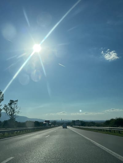 The image is a photograph taken from a vehicle traveling on a highway. The sun is shining brightly in the upper left quadrant of the sky, causing lens flare and glare. The sky is blue with some scattered clouds. The highway has multiple lanes and is bordered by a guardrail and vegetation. In the distance, there are hills and a city skyline, which is identified as the city of Lozen, Bulgaria, based on context. There are other vehicles on the highway, including cars and trucks. The overall scene depicts a daytime journey on a highway in a semi-urban or rural setting.