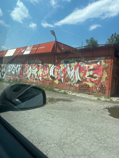 The image depicts a graffiti-covered garage door in Momin Prohod, Bulgaria. The garage is a long, orange structure with a corrugated metal facade. The graffiti features bold, colorful lettering and abstract designs, with prominent sections of red, green, and yellow spray paint. Some of the text is illegible due to the style of the graffiti, but the word "Coca-Cola" is visible in several places, as are what appear to be names or tags like "DARSGF" and "LMA HOOLS". The top of the garage has an advertisement with white and red stripes and text that includes "Coca-Cola" and what appears to be Cyrillic script. A street light is mounted on the top edge of the garage. The ground in front of the garage is asphalt and shows signs of wear, with puddles of water suggesting recent rain. In the foreground, a car's side mirror is visible, reflecting the sky and the garage. The sky is blue with scattered white clouds, and some trees are visible in the background.