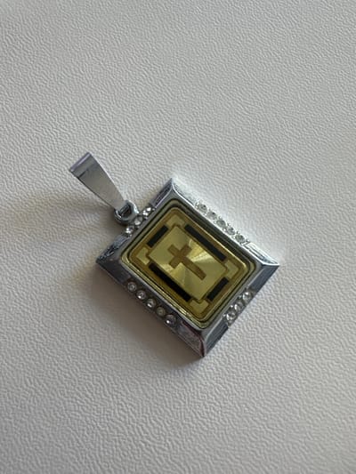The image shows a close-up, top-down view of a pendant lying on a textured, light-colored surface, likely a table or cloth. The pendant is diamond-shaped with a silver-colored metallic frame. The frame is adorned with small, clear stones, possibly diamonds or rhinestones, along its edges. Inside the frame, there is a rectangular design with a gold-colored centerpiece featuring a cross-like symbol in a darker gold hue. Two black rectangular accents are placed on either side of the gold centerpiece within the frame. A silver-colored bail or loop is attached to the bottom point of the pendant, allowing it to be hung. The overall impression is of a piece of jewelry. There is no visible text. The lighting suggests an indoor setting, possibly during the daytime due to the bright illumination. The setting is not explicitly identifiable beyond the pendant and its immediate surface.