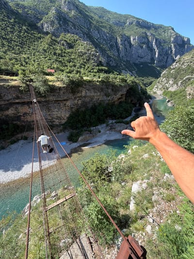 This image, captured in an unknown location in Montenegro, presents a high-angle view looking down into a deep, verdant river canyon on a clear, sunny day.

In the immediate foreground, a light-skinned male's right arm and hand are prominently positioned, with the index finger extended and pointing into the canyon, while the thumb is also extended, forming a "shaka" or "hang loose" gesture. The arm is tan with visible hair.

Below the pointing hand, a rustic, potentially aged, suspension bridge constructed of metal cables and a grid-like mesh walkway spans the canyon. The metal components of the bridge exhibit signs of surface rust, and some wooden planks are integrated into the walkway. Two individuals and a dog are seen traversing the bridge. The closest person, with their back to the camera, wears a white t-shirt, dark shorts, and white sneakers, and appears to be carrying a black backpack or bag. A medium-sized black dog walks directly behind this person. Further along the bridge, a second person in a light-colored top and dark bottoms also walks away from the camera.

The bridge descends towards a pebbly riverbank with a clear, turquoise-green river flowing through the bottom of the canyon. The river is relatively wide with shallow areas visible. On the far bank, a small building with a red tile roof is nestled among the dense green trees, partially obscured by the foliage. A dark, possibly wooden, structure or platform is also visible near the far end of the bridge's connection point to the land. A blue tarp or similar object is discernible on the riverbank below the bridge.

The canyon walls are steep and mountainous, covered extensively with lush green trees and varied vegetation. Exposed grey and light brown rock faces are visible through the foliage, particularly on the larger mountain in the background. The sky above is a clear, bright blue, indicating fair weather. Strong sunlight casts distinct shadows across the landscape. The suspension bridge's rusty metal anchor points are visible in the bottom right corner of the foreground.